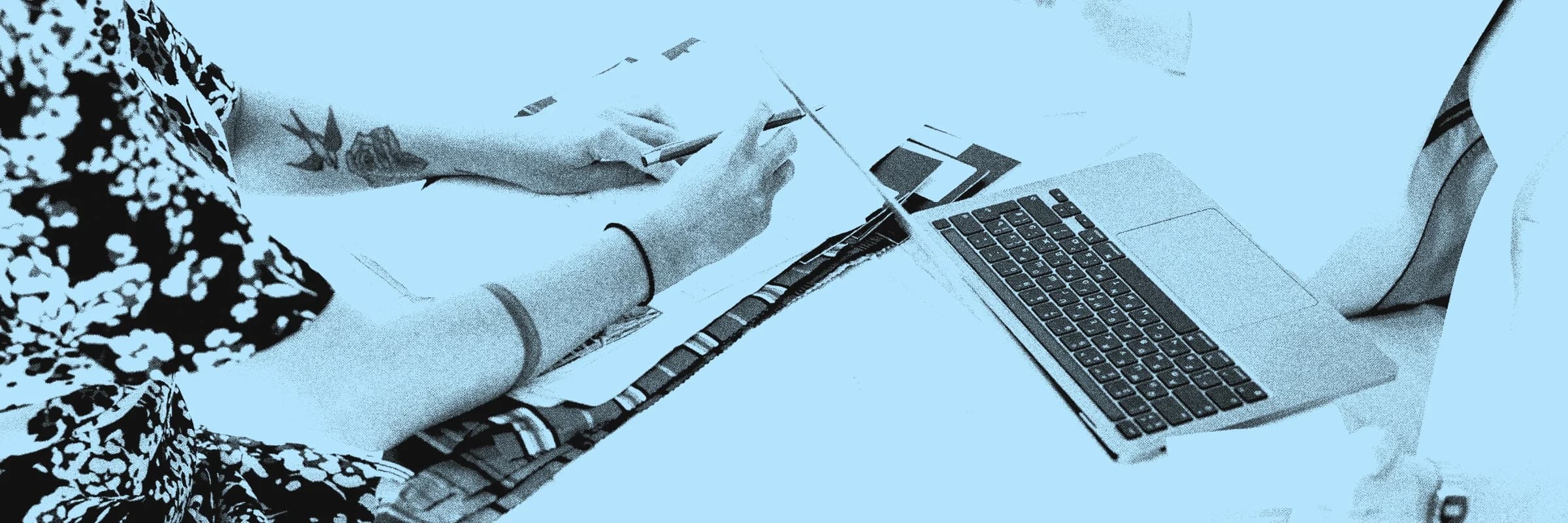 Duas pessoas a uma mesa com laptop, desenhos técnicos, retalhos de tecido e post-its, vistas sem os rostos no enquadramento.